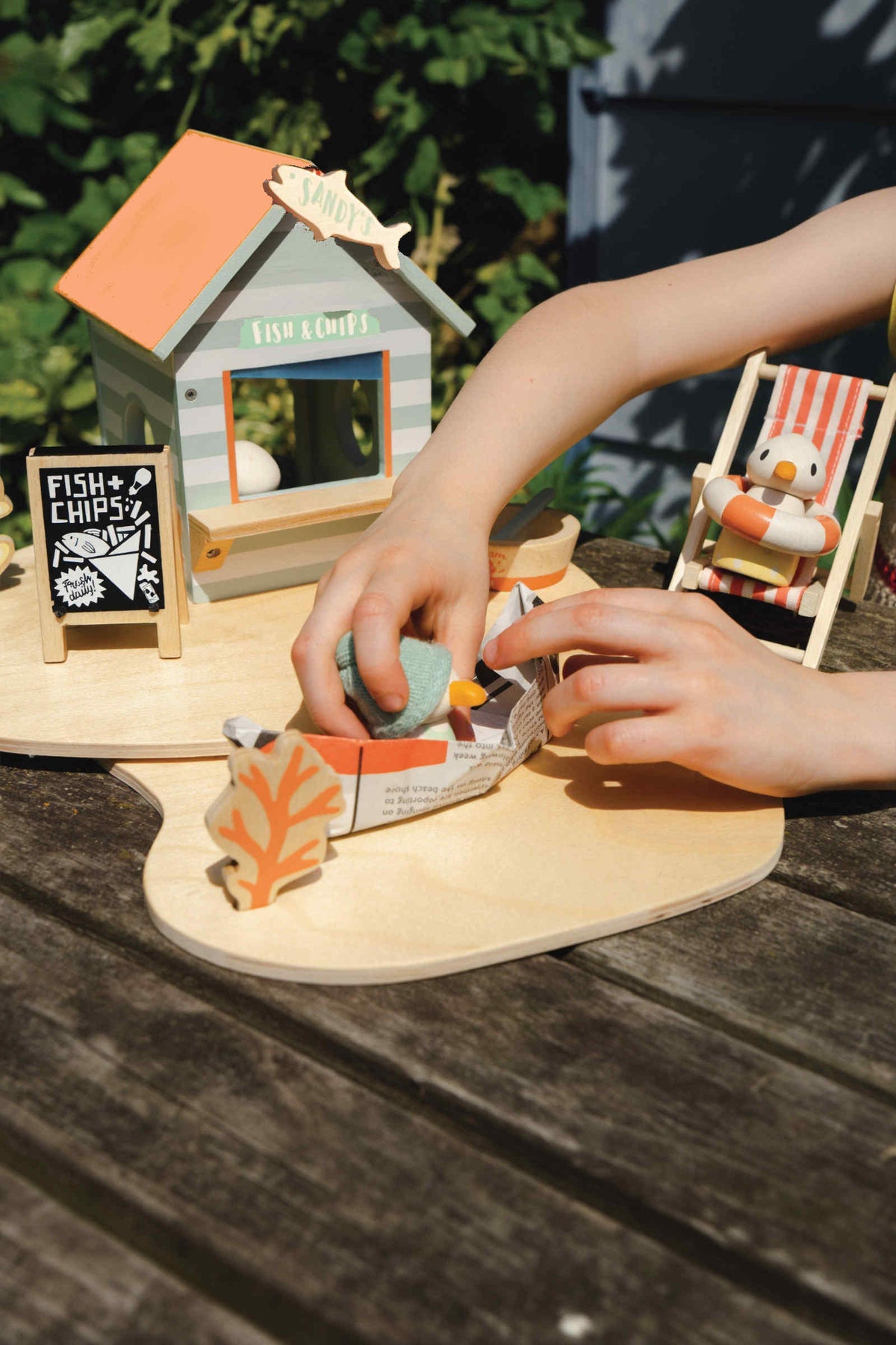 A child plays with Tender Leafs Sandys Beach Hut toy set, featuring a wooden fish and chips stall and colorful.