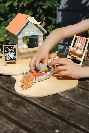 A child plays with Tender Leafs Sandys Beach Hut toy set, featuring a wooden fish and chips stall and colorful.