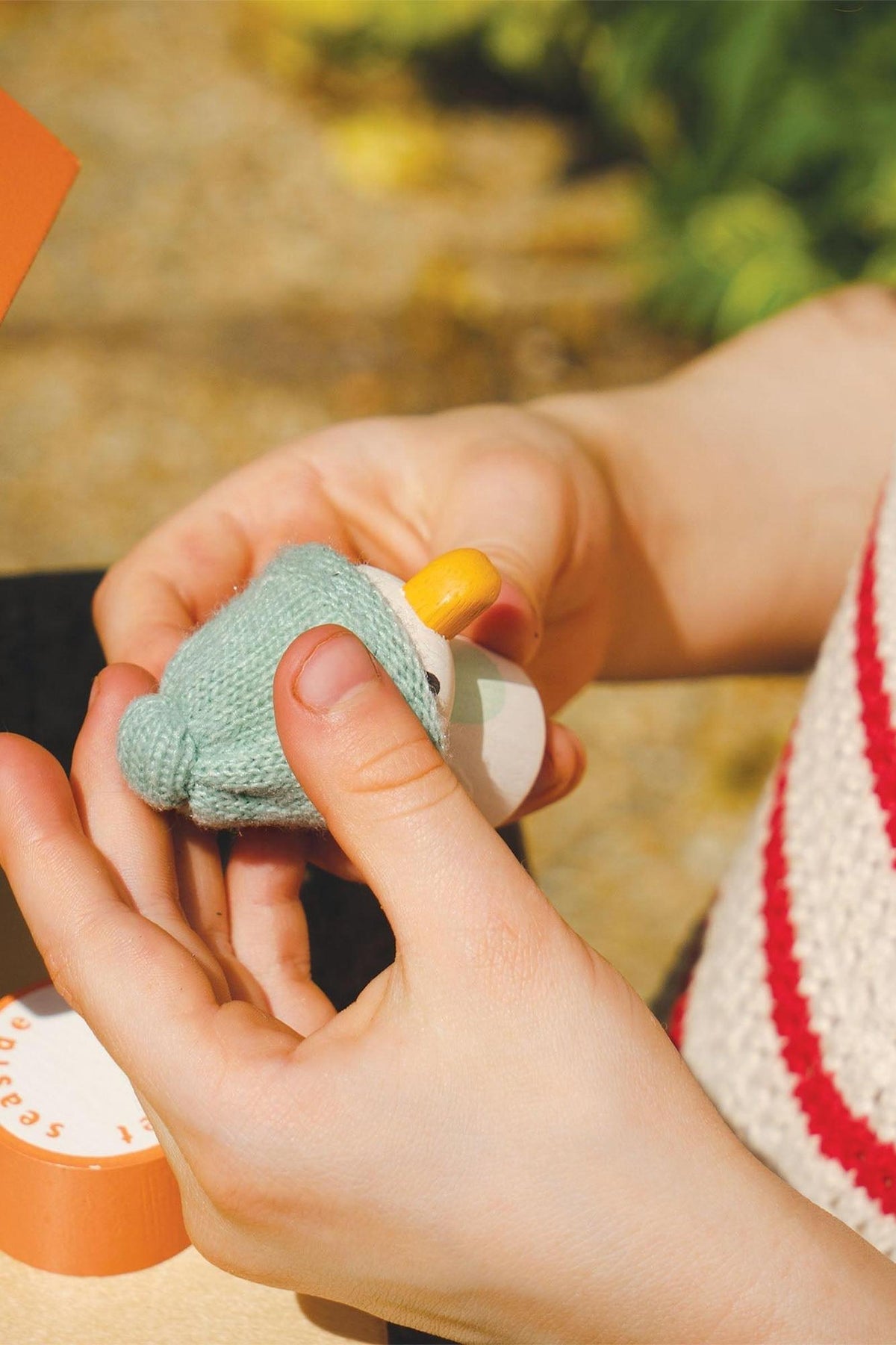 A child holds a small, colorful toy resembling a duck, crafted from soft materials. The setting features natural outdoor lighting.