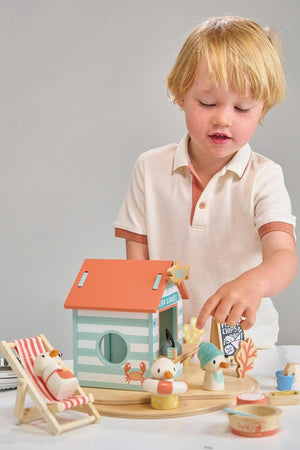 A young child plays with Tender Leaf Wooden Toys Sandys Beach Hut, featuring colorful wooden figures and a cozy beach.