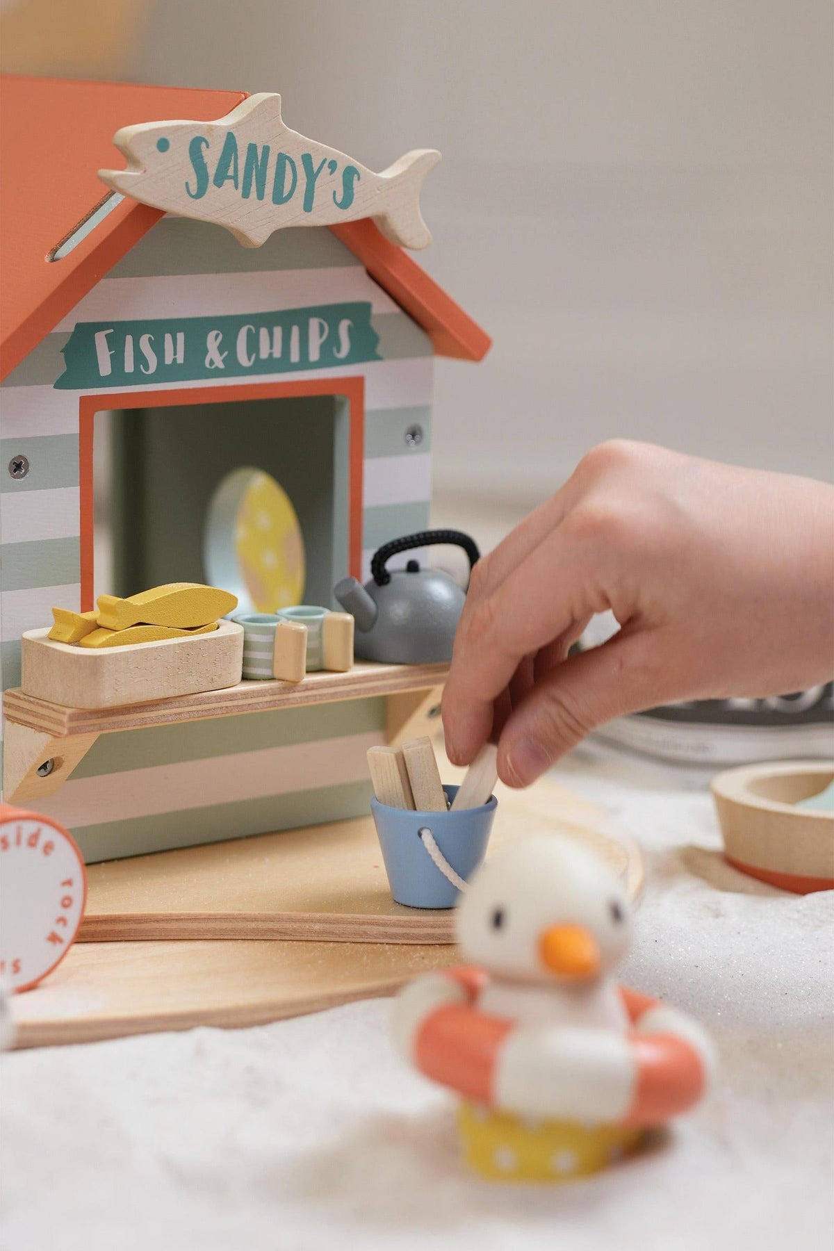 A child’s hand places a wooden piece into a blue bucket beside Sandys Fish and Chips stall, featuring pastel colors.