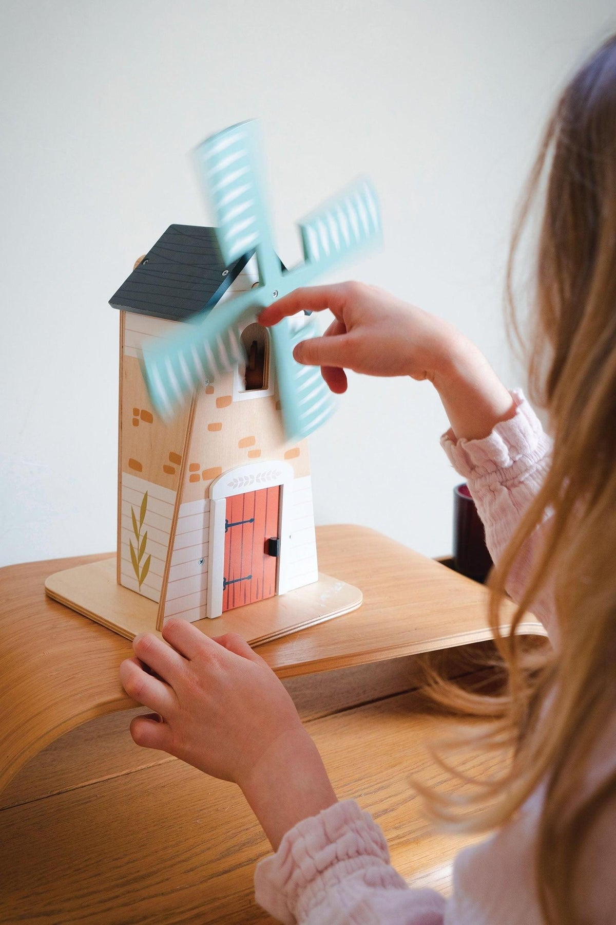 A child gently spins the blue blades of a wooden windmill playset, featuring a colorful two-story house with a red door and decorative details, promoting imaginative play.