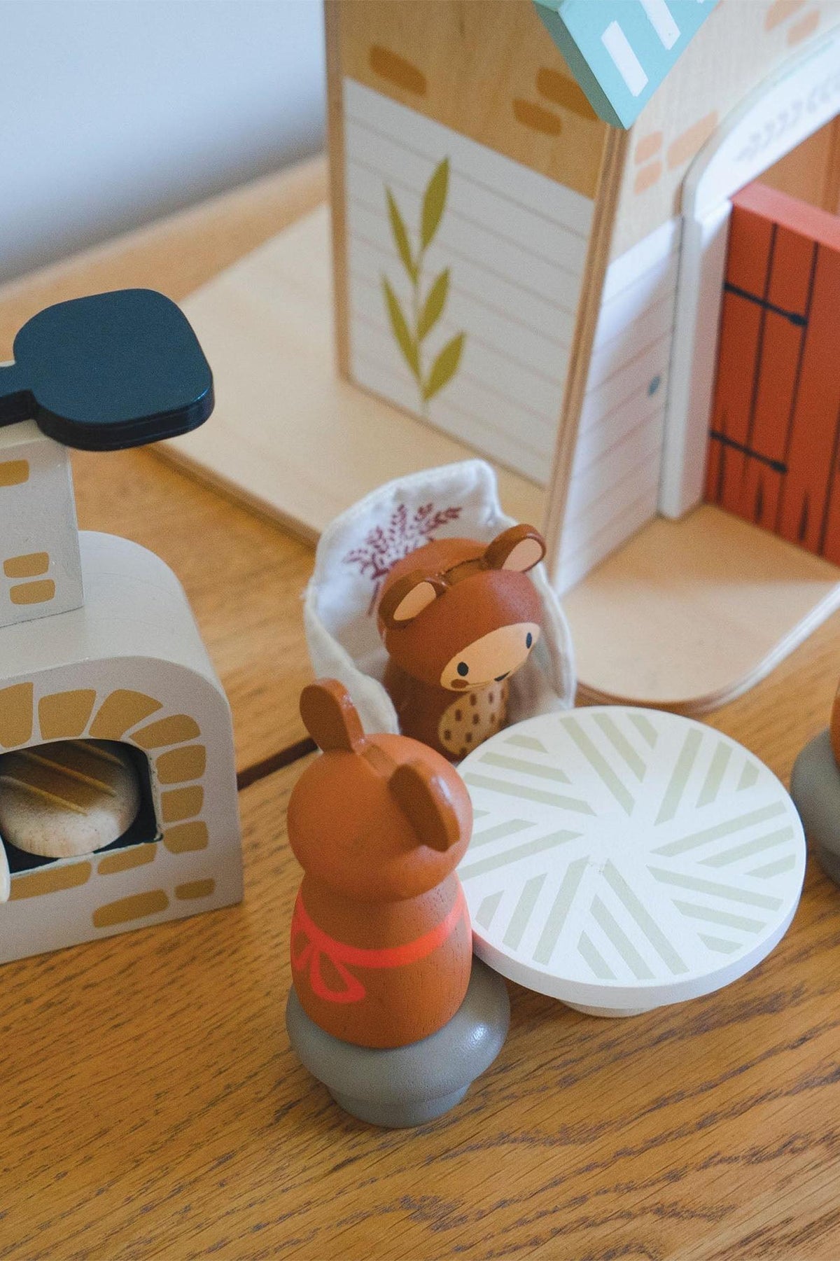 Two wooden field mice figures, one with a brown coat and red scarf, stand beside a round patterned table in a cozy wooden playset.
