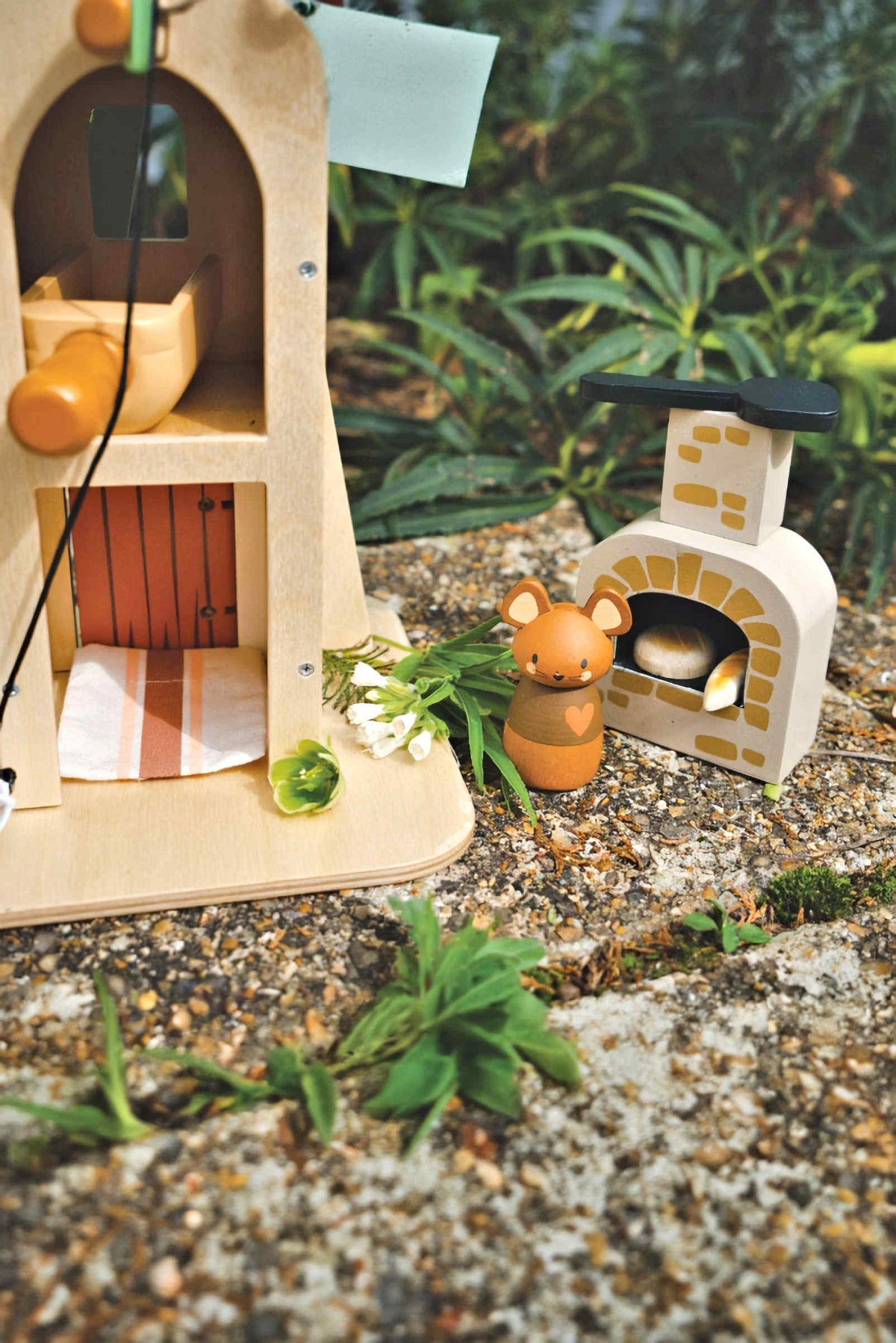 A wooden playset features a two-story design with a revolving windmill and a cozy interior, accompanied by a charming bear figurine next to a stone fireplace, surrounded by greenery.