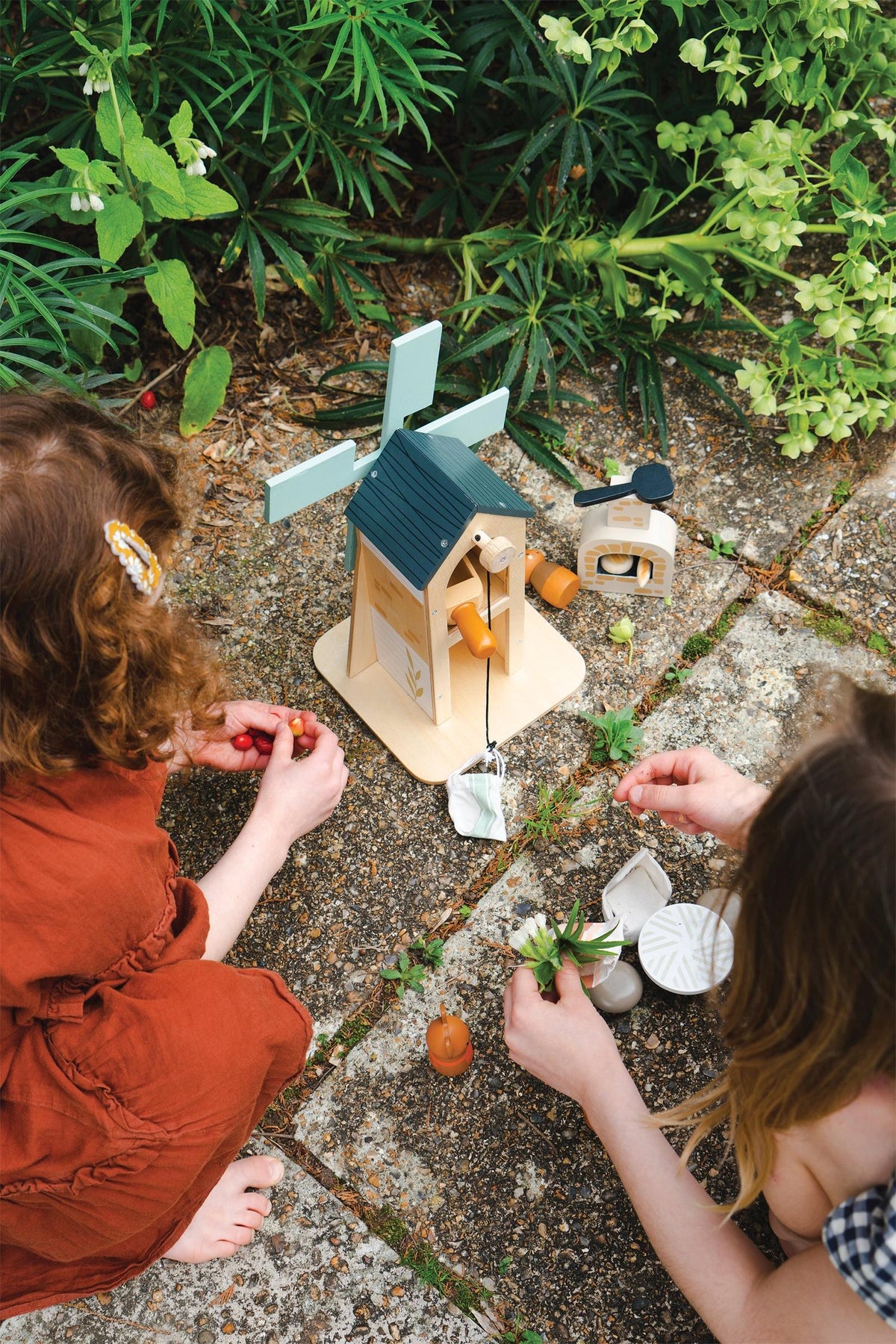 Two children play with the Tender Leaf Wooden Toys Penny Windmill, a wooden playset featuring a green-roofed windmill and charming accessories, surrounded by lush greenery on a stone patio.