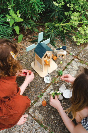 Two children play with the Tender Leaf Wooden Toys Penny Windmill, a wooden playset featuring a green-roofed windmill and charming accessories, surrounded by lush greenery on a stone patio.
