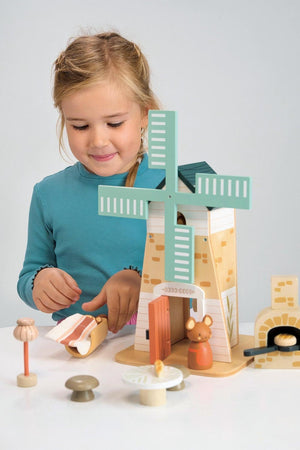 A young girl interacts with the Tender Leaf Wooden Toys Penny Windmill playset, featuring a charming two-story wooden structure with a green windmill and adorable field mouse figures.
