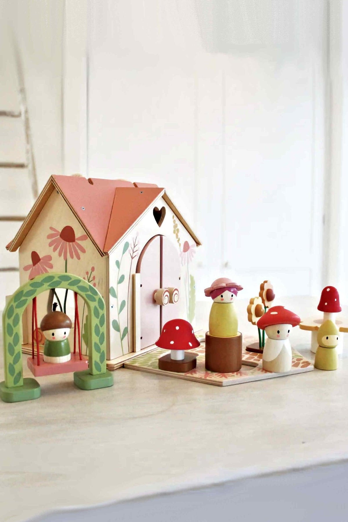 A charming wooden playhouse with a rose-colored roof, decorated with flowers, accompanied by colorful mushroom figurines on a light surface.
