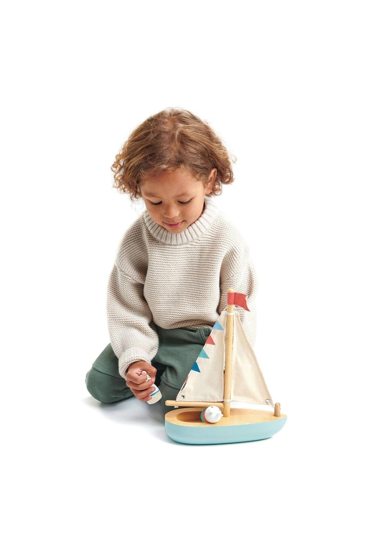 A child in a cozy sweater plays with a Tender Leaf wooden sailboat, featuring fabric sails and colorful bunting.