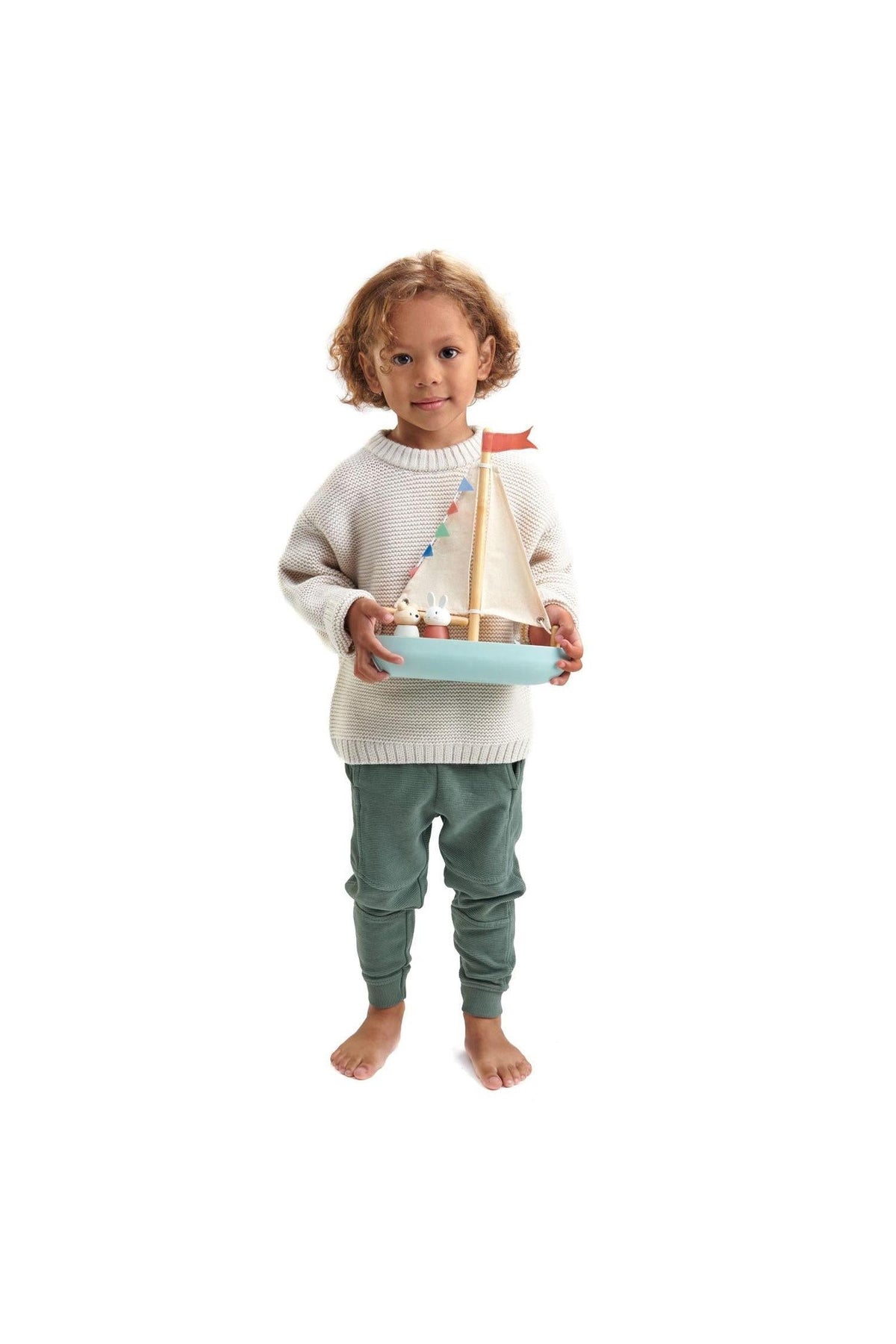 A child in a light sweater and green pants holds a wooden sailboat with fabric sails and decorative bunting.