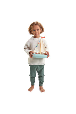 A child in a light sweater and green pants holds a wooden sailboat with fabric sails and decorative bunting.