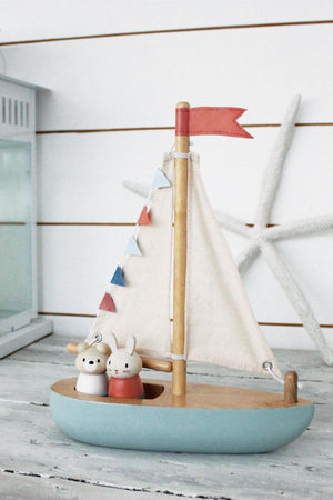 A solid wood sailboat features two charming animal figures, with fabric sails and colorful bunting, set against a coastal backdrop.