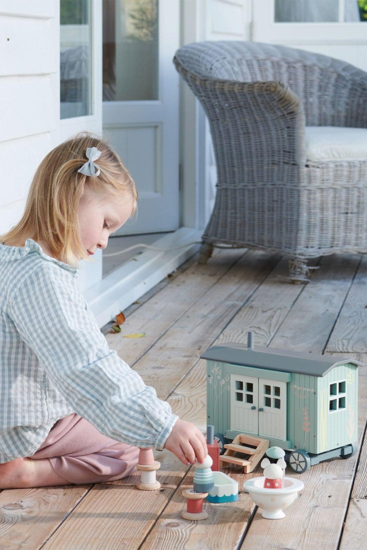 A young girl sits on a wooden porch, playing with the Tender Leaf Wooden Toys Secret Meadow Shepherds Hut. The toy features soft colors and imaginative details, encouraging creative play.