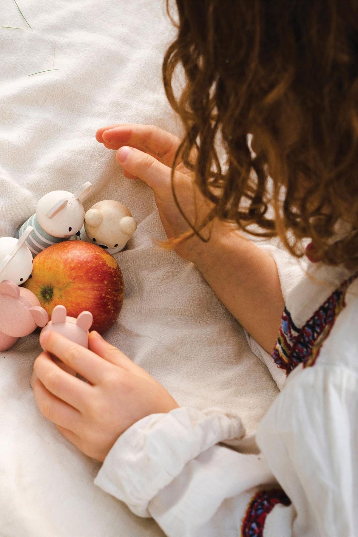 A child with curly hair gently plays with small wooden mice toys and a red apple on a white blanket. The scene captures a cozy and imaginative playtime atmosphere.