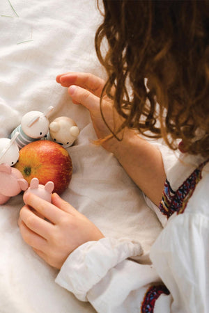 A child with curly hair gently plays with small wooden mice toys and a red apple on a white blanket. The scene captures a cozy and imaginative playtime atmosphere.