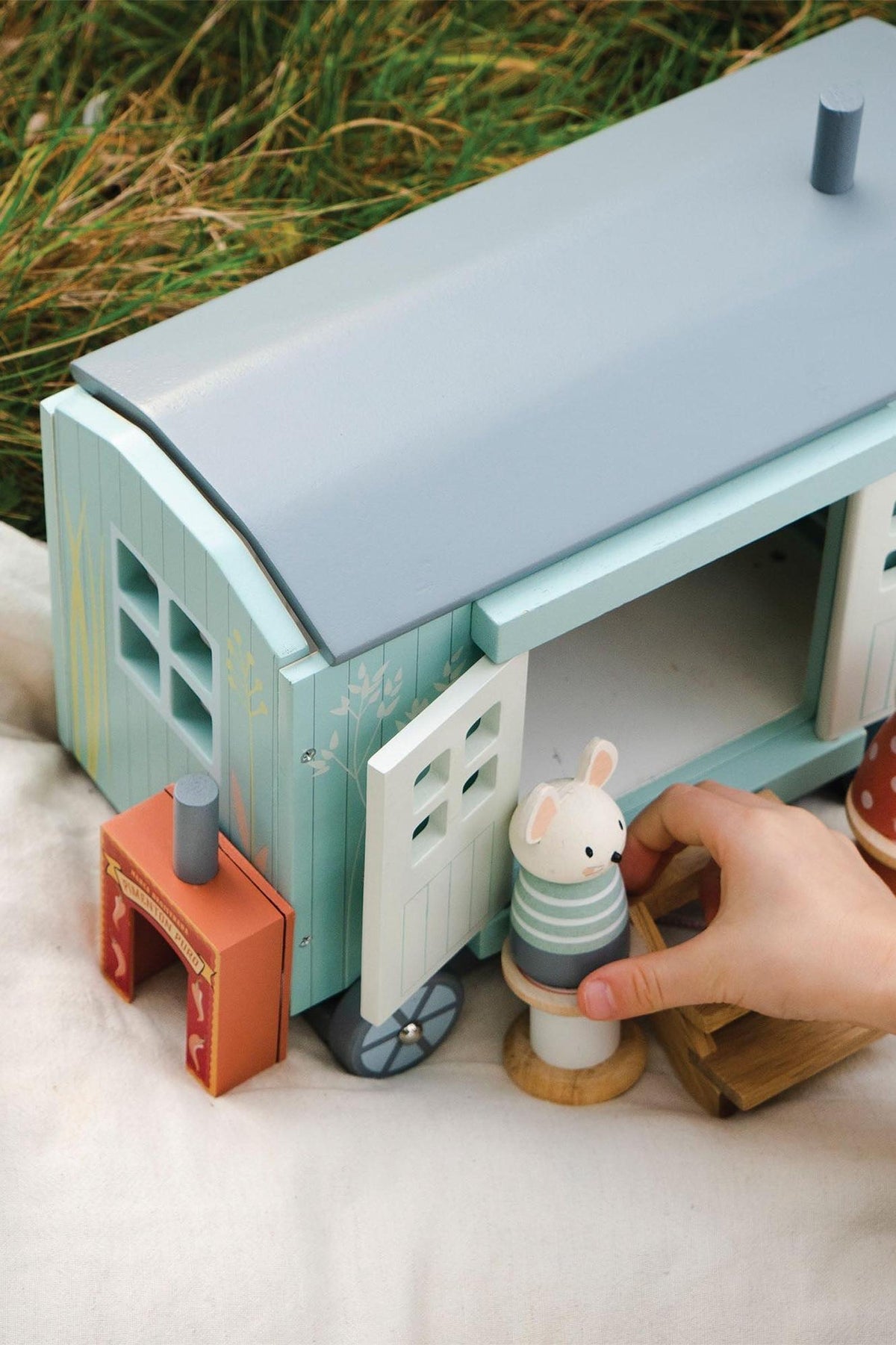 A child’s hand reaches for a wooden mouse figure next to a beautifully crafted pastel green and blue shepherds hut toy, set on soft grass.