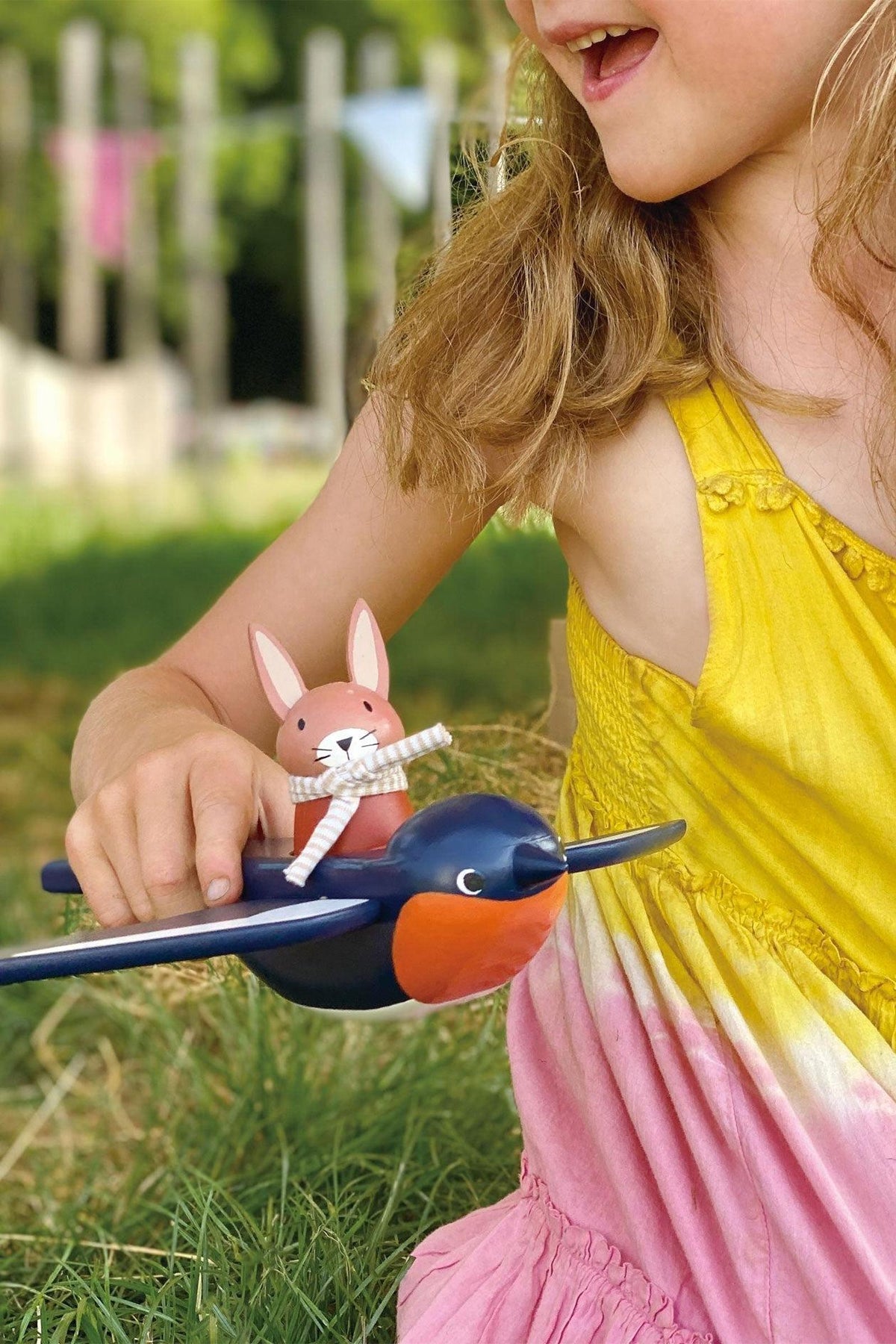 A child in a yellow dress holds a wooden bunny toy on a colorful bird toy, surrounded by grass.