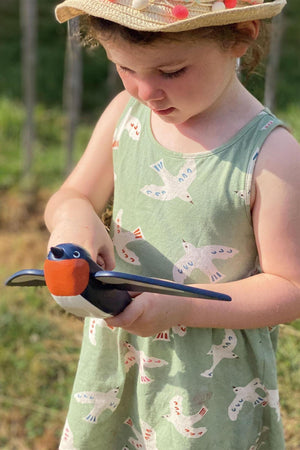 A young child in a bird-patterned dress holds a wooden toy bird, focused on play in an outdoor setting.