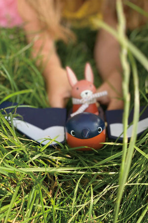 A child plays in grass, holding a wooden bunny toy alongside a colorful bird toy from the Tender Leaf collection.