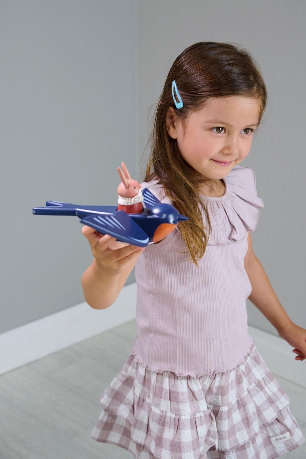 A young girl in a light purple top holds a blue bird toy with a small bunny figure, smiling happily.