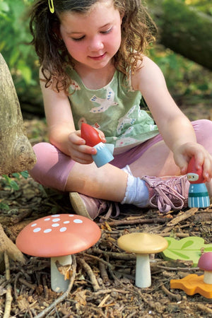 A young girl plays on the forest floor, holding a wooden gnome toy near colorful toadstools. She wears a light green top with bird patterns, surrounded by natural foliage.