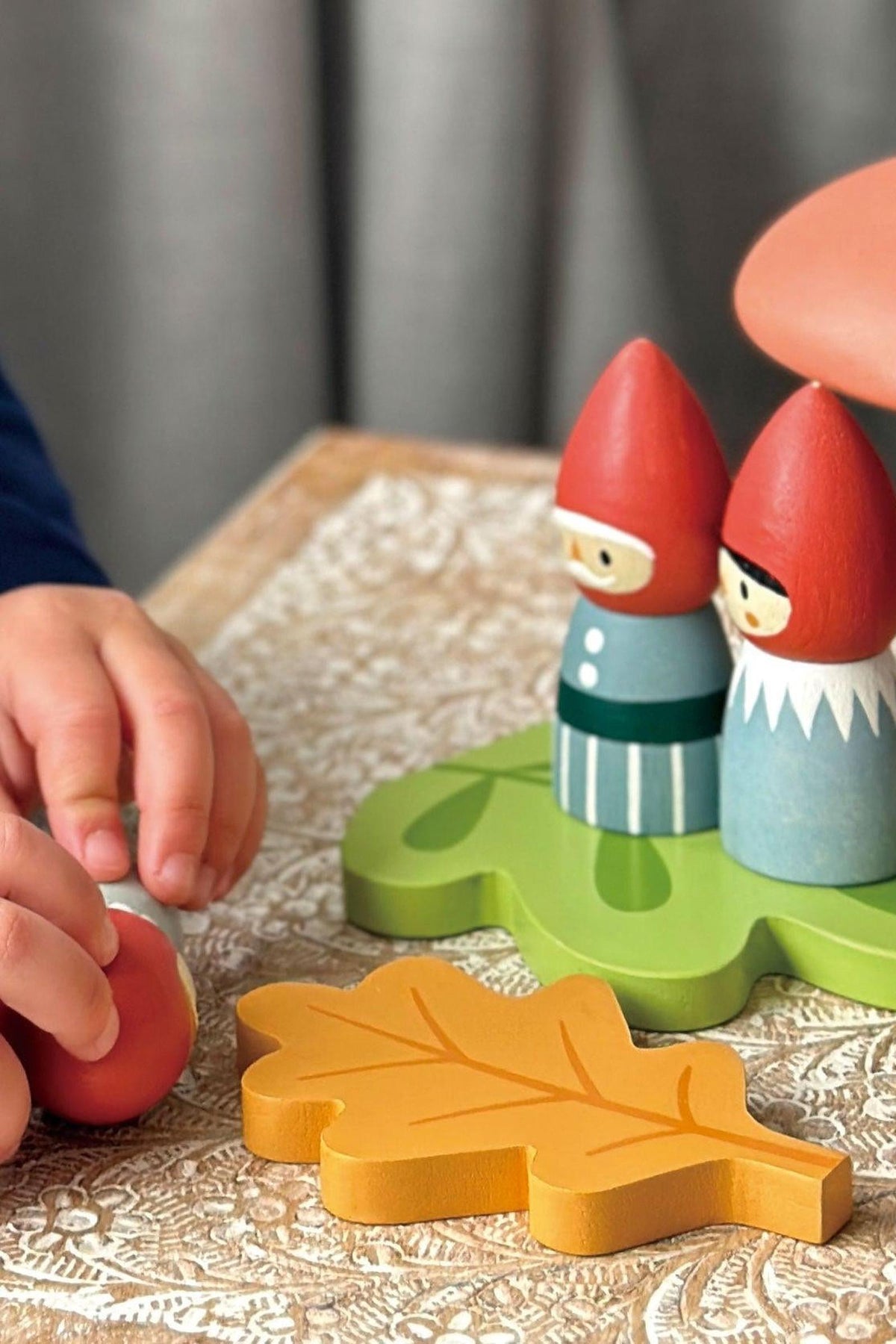 A child’s hand reaches for a small red ball near two wooden gnomes, brightly painted with red hats.