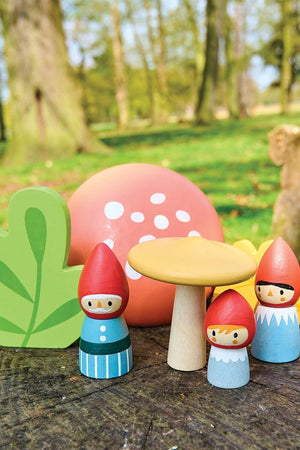 A colorful wooden gnome family set features three gnomes with red hats, a yellow mushroom, and a green leaf in a natural outdoor setting.