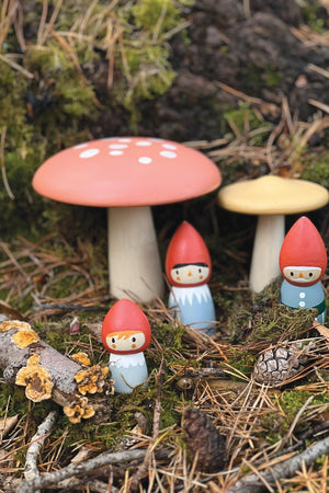 Three colorful wooden gnomes stand among pine needles and moss near oversized toadstools. The gnomes feature red hats and blue and white outfits, highlighting their playful and whimsical design.