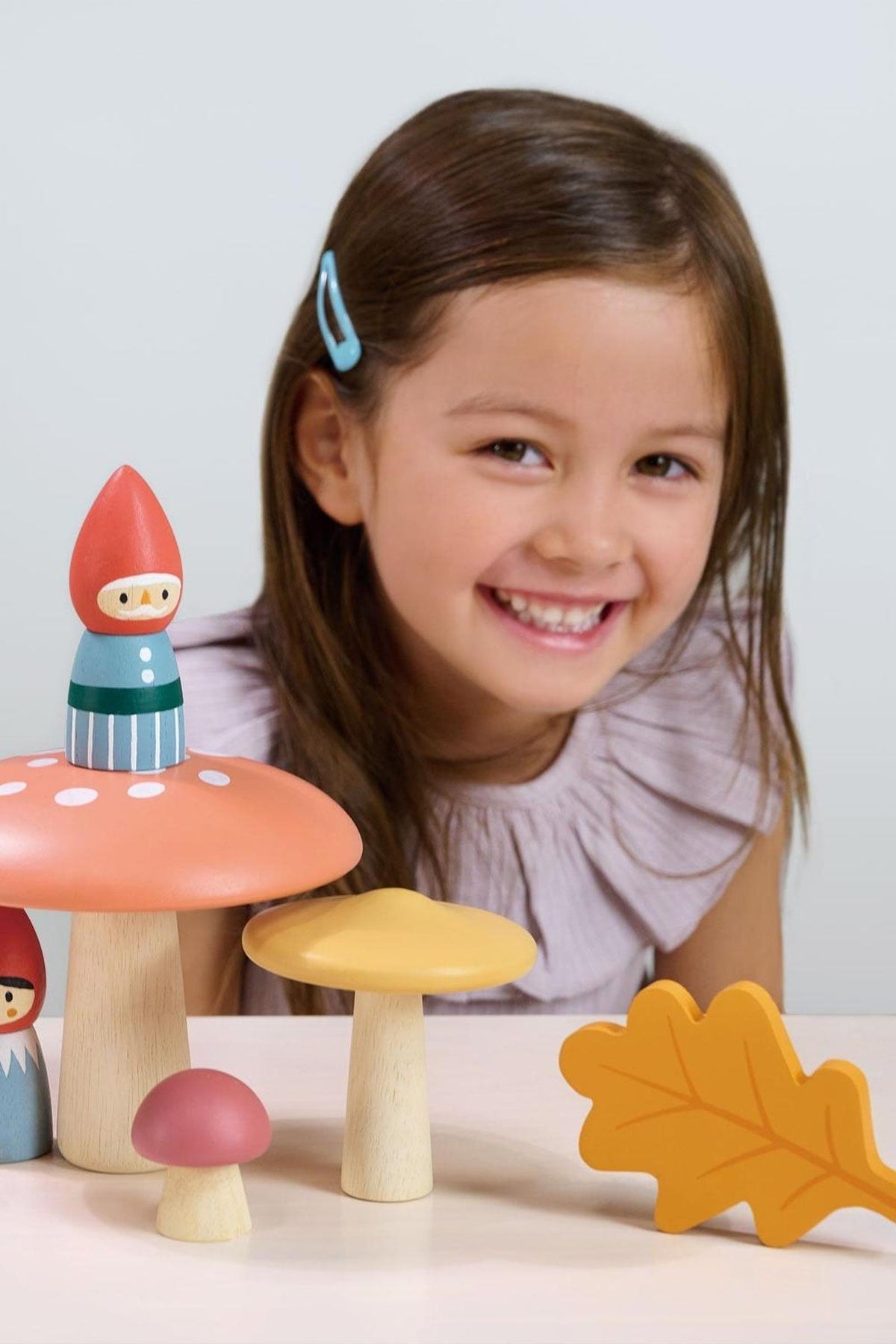 A smiling girl with long brown hair wears a pink top while playing with colorful wooden gnome figures and mushrooms on a light surface, showcasing the Tender Leaf Wooden Toys.
