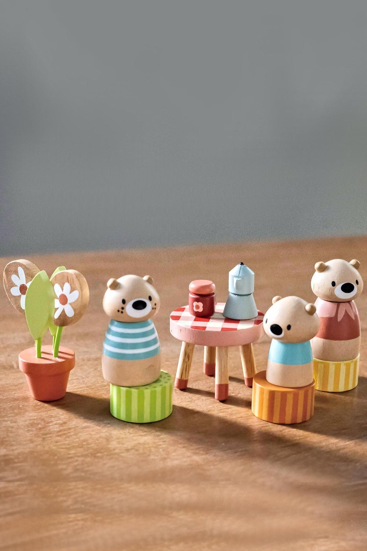 A set of colorful wooden bear figurines, featuring striped shirts, sits beside a small table with a checkered tablecloth, a candle, and a flower pot, on a wooden surface.