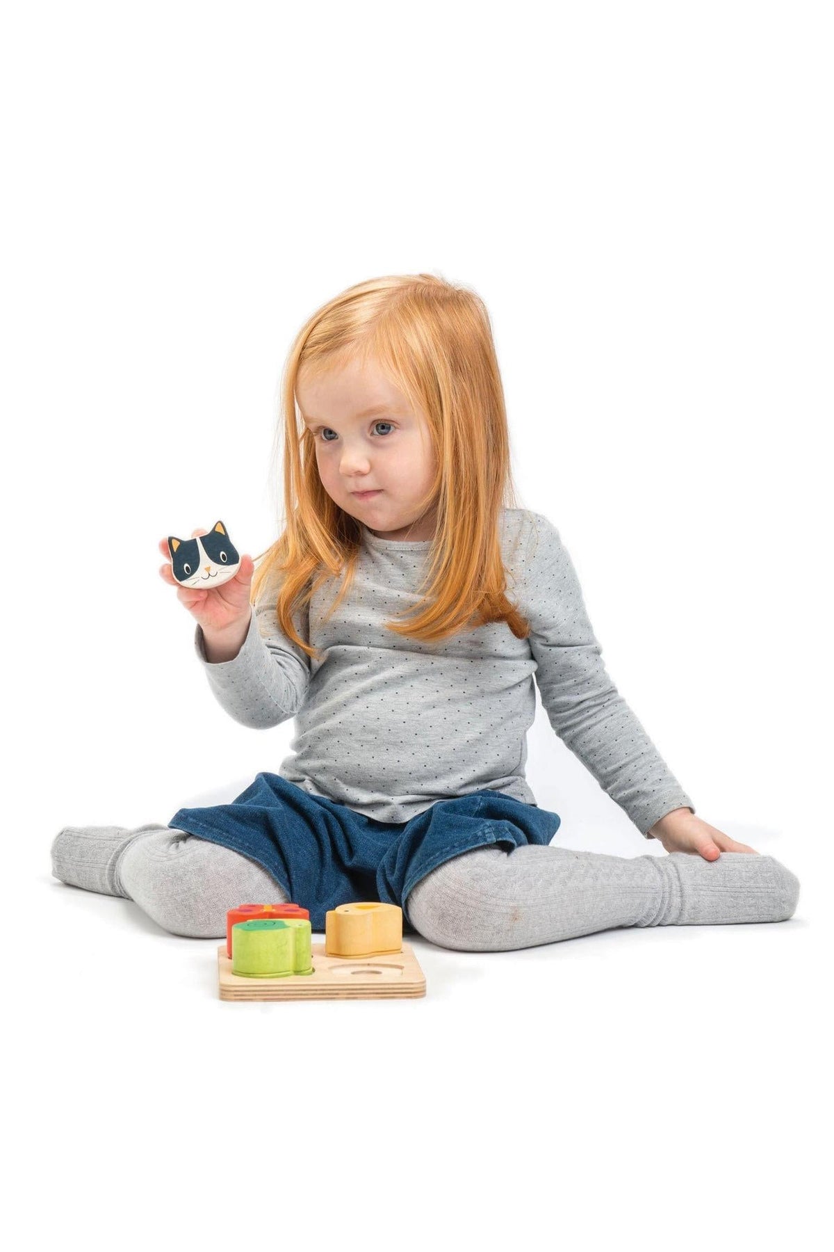 A toddler in a grey outfit holds a cat-shaped wooden piece, with colorful blocks on the floor.