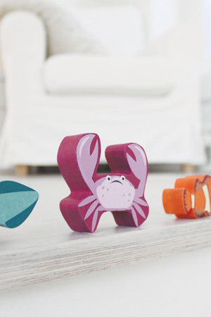 A vibrant pink crab toy stands among green and orange sea creature figures on a light wooden surface.