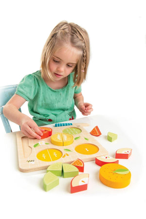 A young girl in a green shirt plays with a colorful wooden fruit fraction puzzle, featuring slices of citrus fruits on a wooden board.