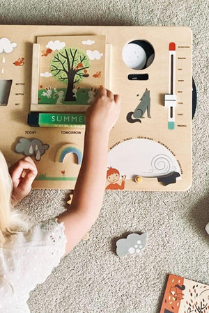 A child interacts with the Tender Leaf Wooden Toys Weather Watch, featuring colorful illustrations of seasons, weather symbols, and a tree, on a light wooden board.