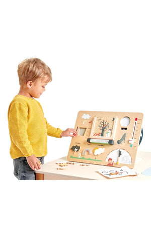 A young child interacts with a wooden weather station toy, featuring colorful illustrations of seasons and weather elements. The toy is made from sustainable materials, promoting educational play.