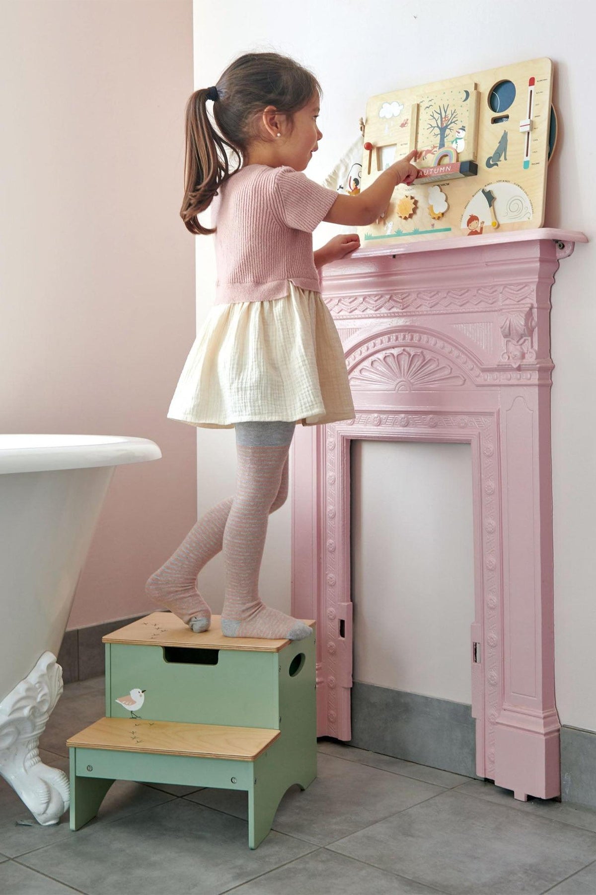 A young girl stands on a green wooden step stool, interacting with a colorful weather station mounted on a pink decorative wall.