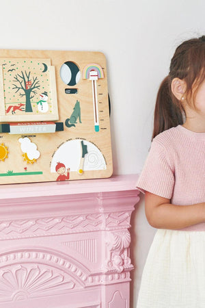 A child stands beside a colorful wooden weather station featuring illustrations of seasonal elements, including a snowman and animals, against a pink mantel. The educational toy promotes interactive learning.