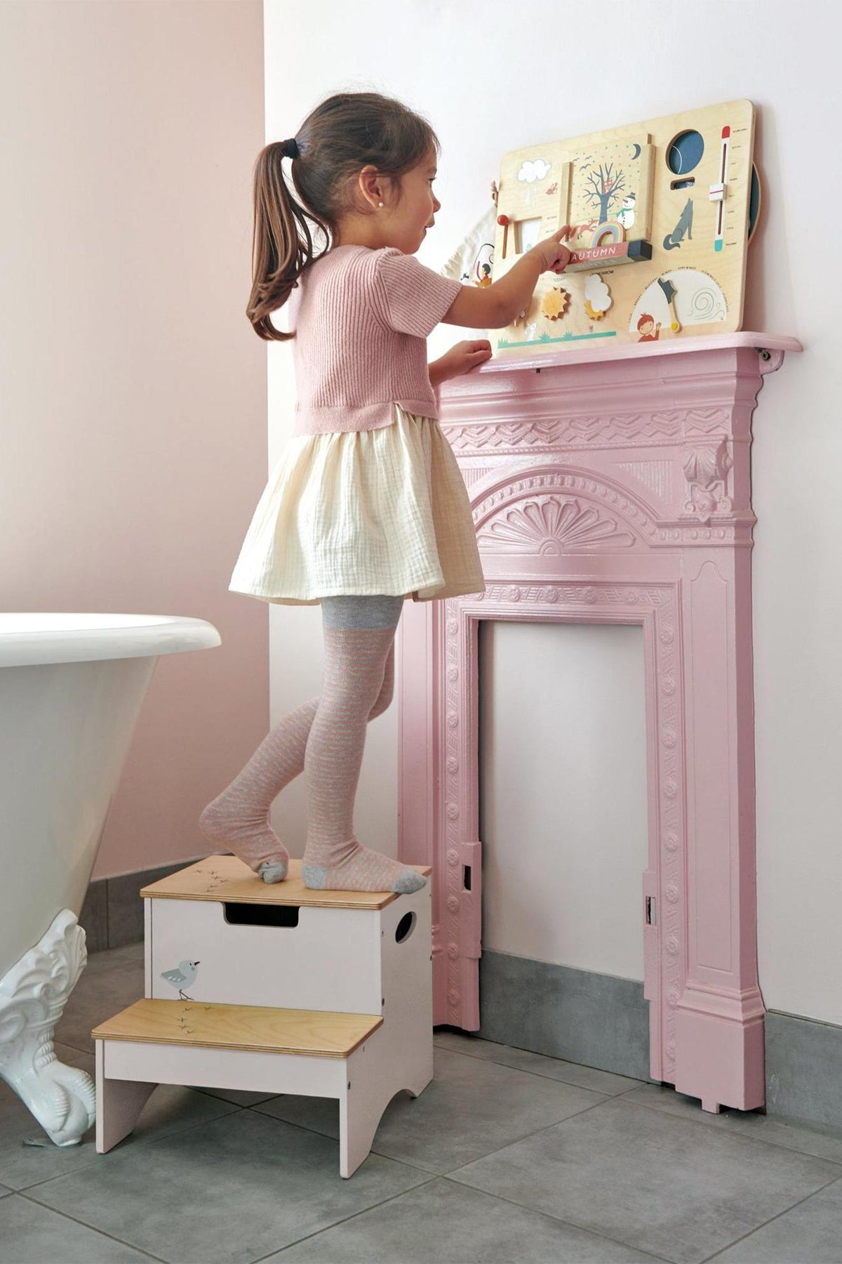 A young girl stands on a wooden, two-step stool reaching for a colorful wall-mounted activity board. The stool features a hidden storage compartment and is designed for toddlers.