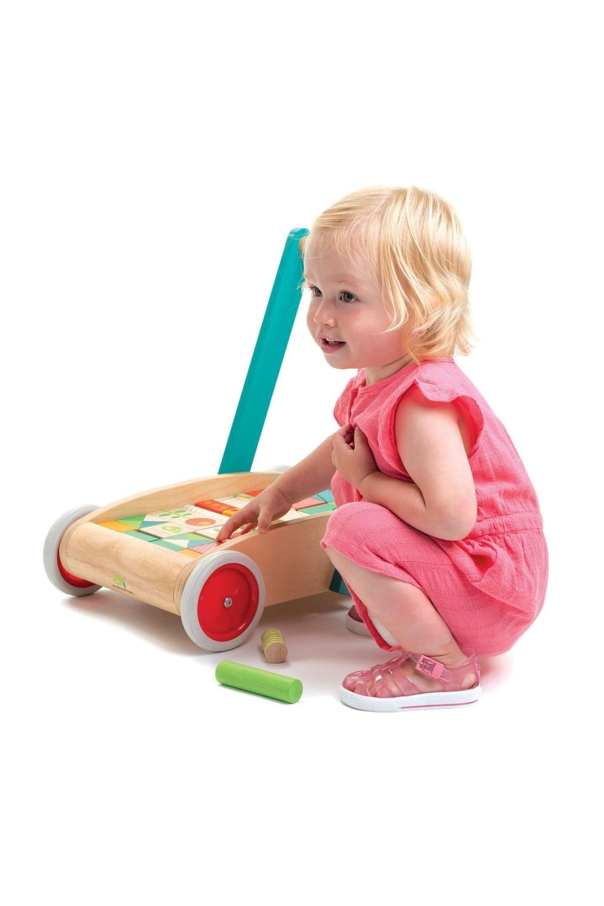 A toddler in a pink outfit crouches beside a Tender Leaf wooden baby block walker, which features a vibrant handle and colorful blocks for playful construction.