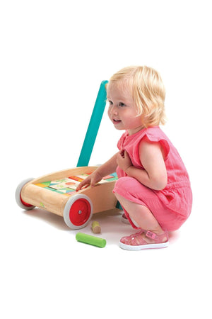 A toddler in a pink outfit crouches beside a Tender Leaf wooden baby block walker, which features a vibrant handle and colorful blocks for playful construction.