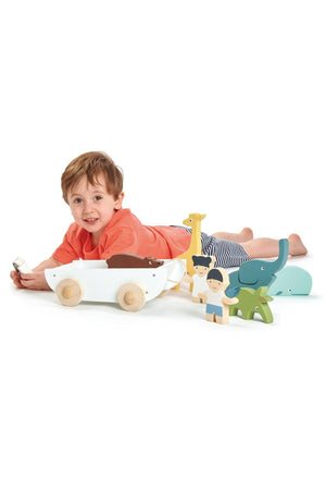 A child lies on the floor, playing with a wooden toy boat and various colorful animal figures made from sustainable.