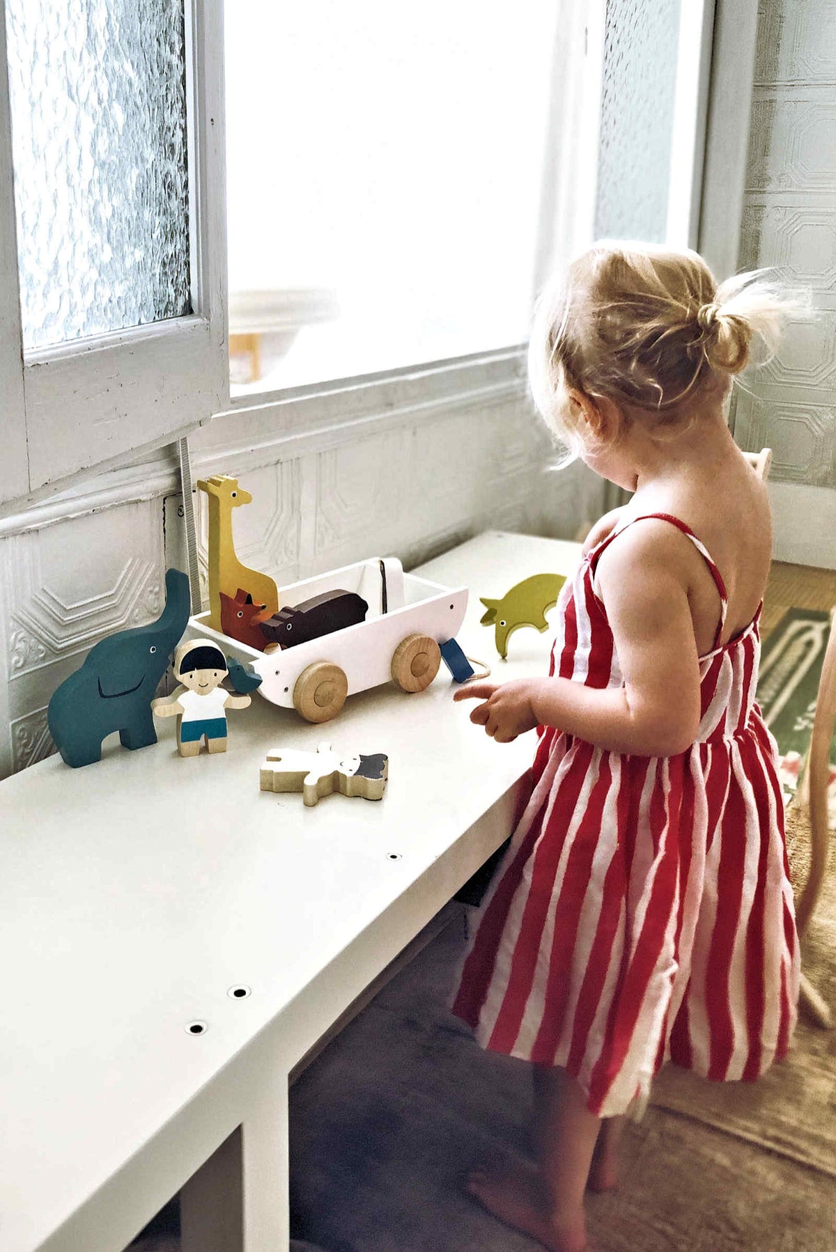 A young child in a striped dress plays with colorful wooden animal toys and a toy cart on a white.