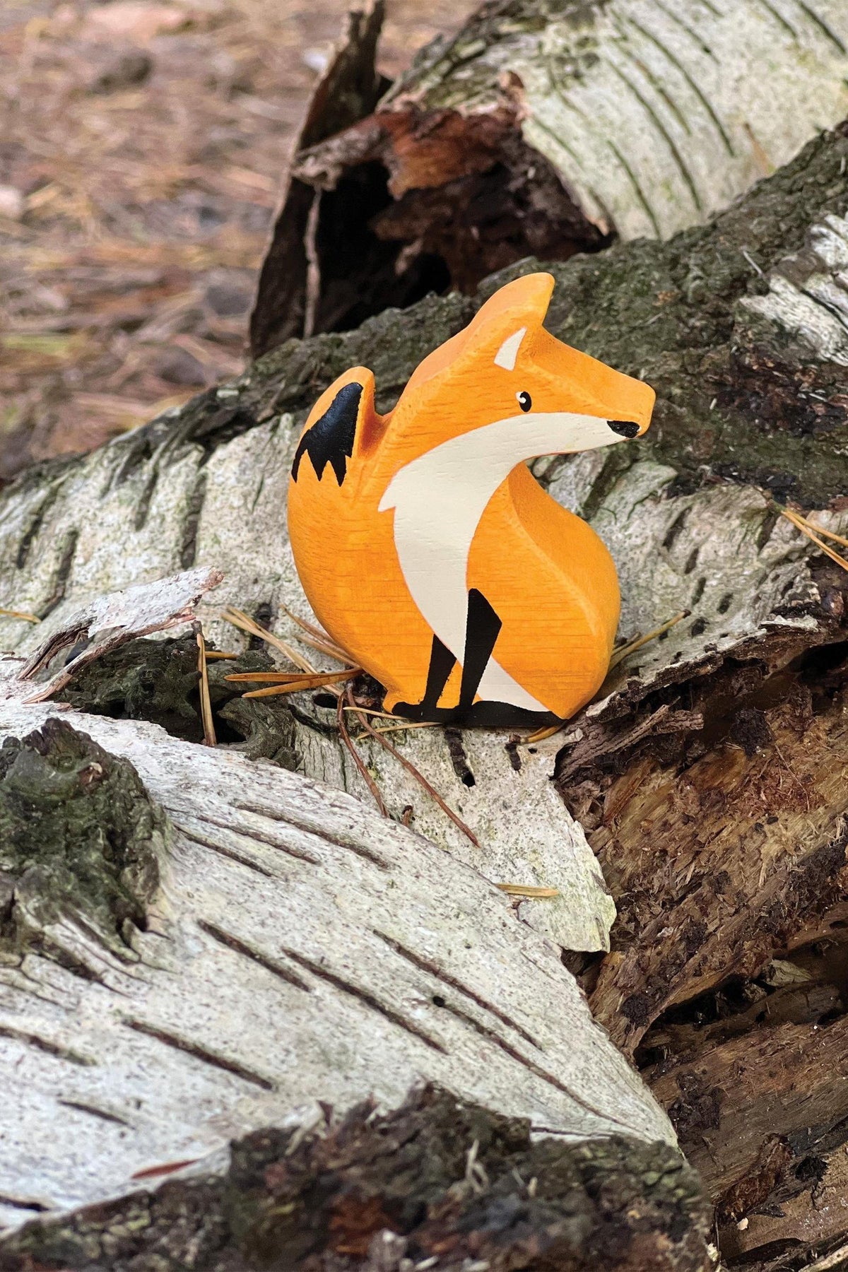 A brightly colored wooden fox figurine sits on a bed of bark and birch tree bark, showcasing its handcrafted details and vibrant orange hue, ideal for imaginative play.