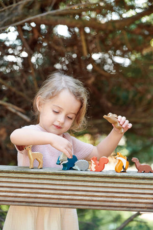 A young girl plays with a set of colorful wooden animal toys, including a fox and deer, arranged on a wooden surface amidst a natural outdoor setting.