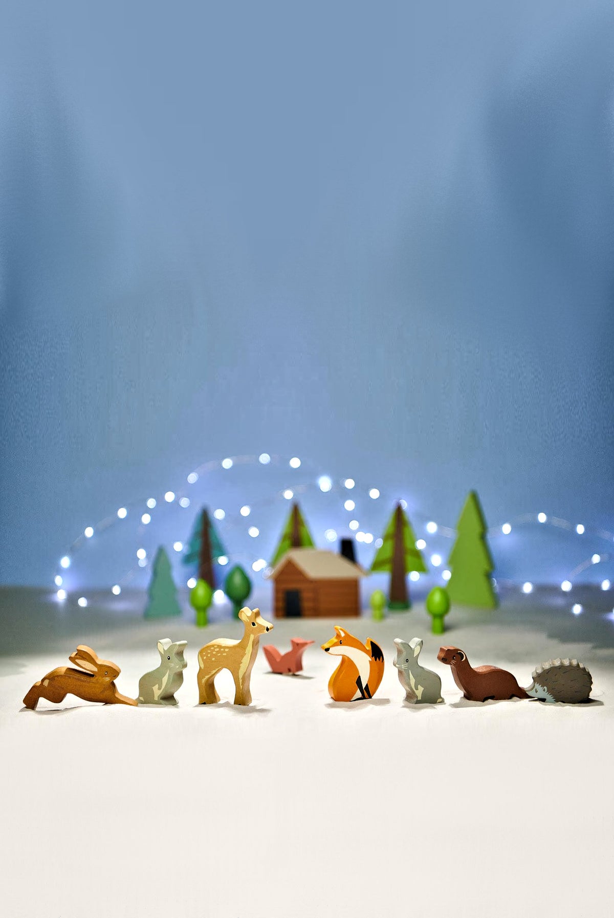 A colorful display of wooden woodland animal figurines, including a fox, deer, and hedgehog, arranged in front of a small cabin and softly lit trees against a blue background.