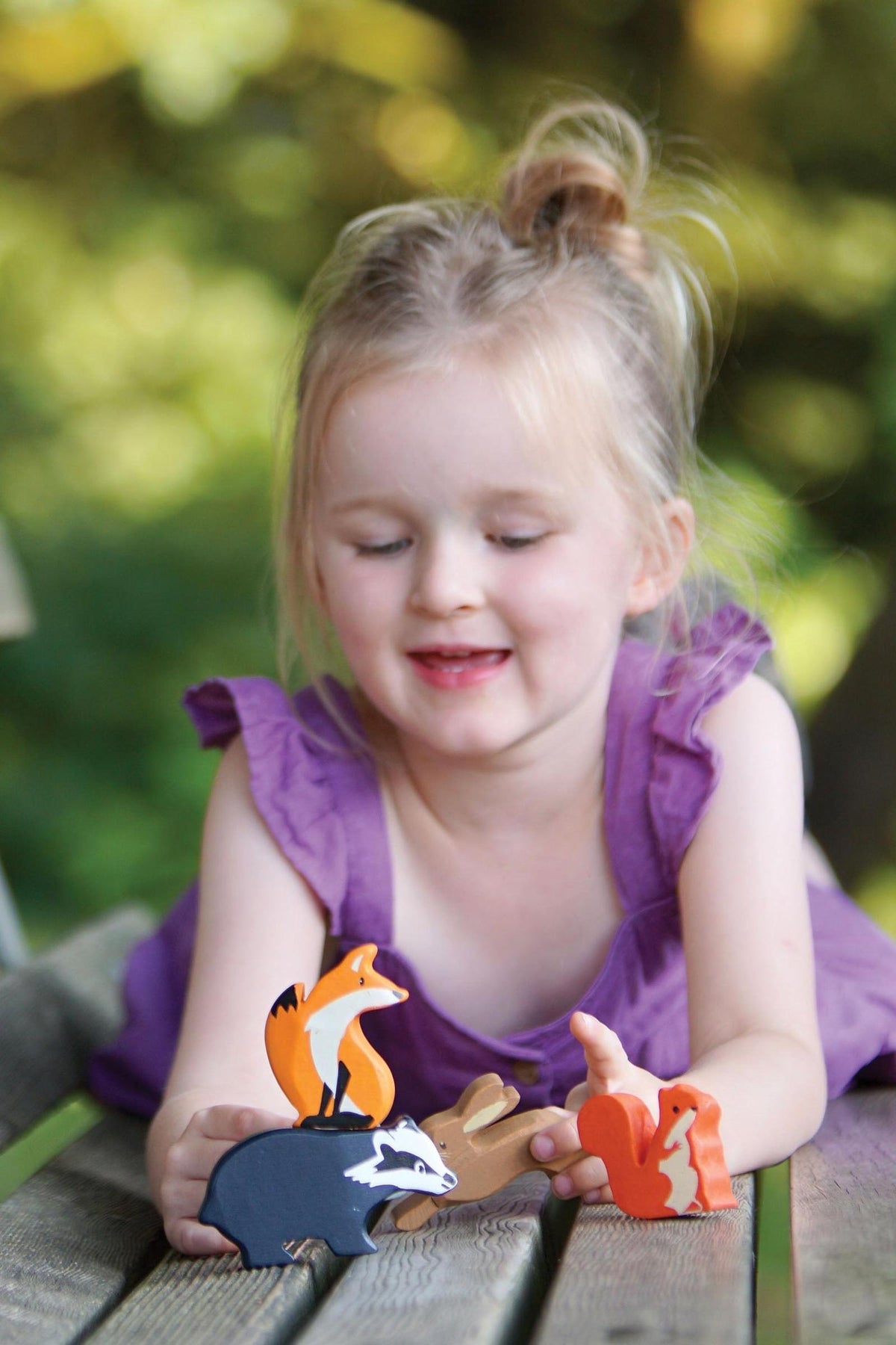 A young girl in a purple dress smiles while playing with colorful wooden animal figures, including a fox, badger, and squirrel, on a wooden table outdoors.
