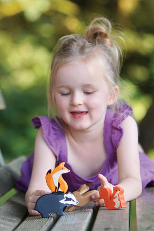 A young girl in a purple dress smiles while playing with colorful wooden animal figures, including a fox, badger, and squirrel, on a wooden table outdoors.
