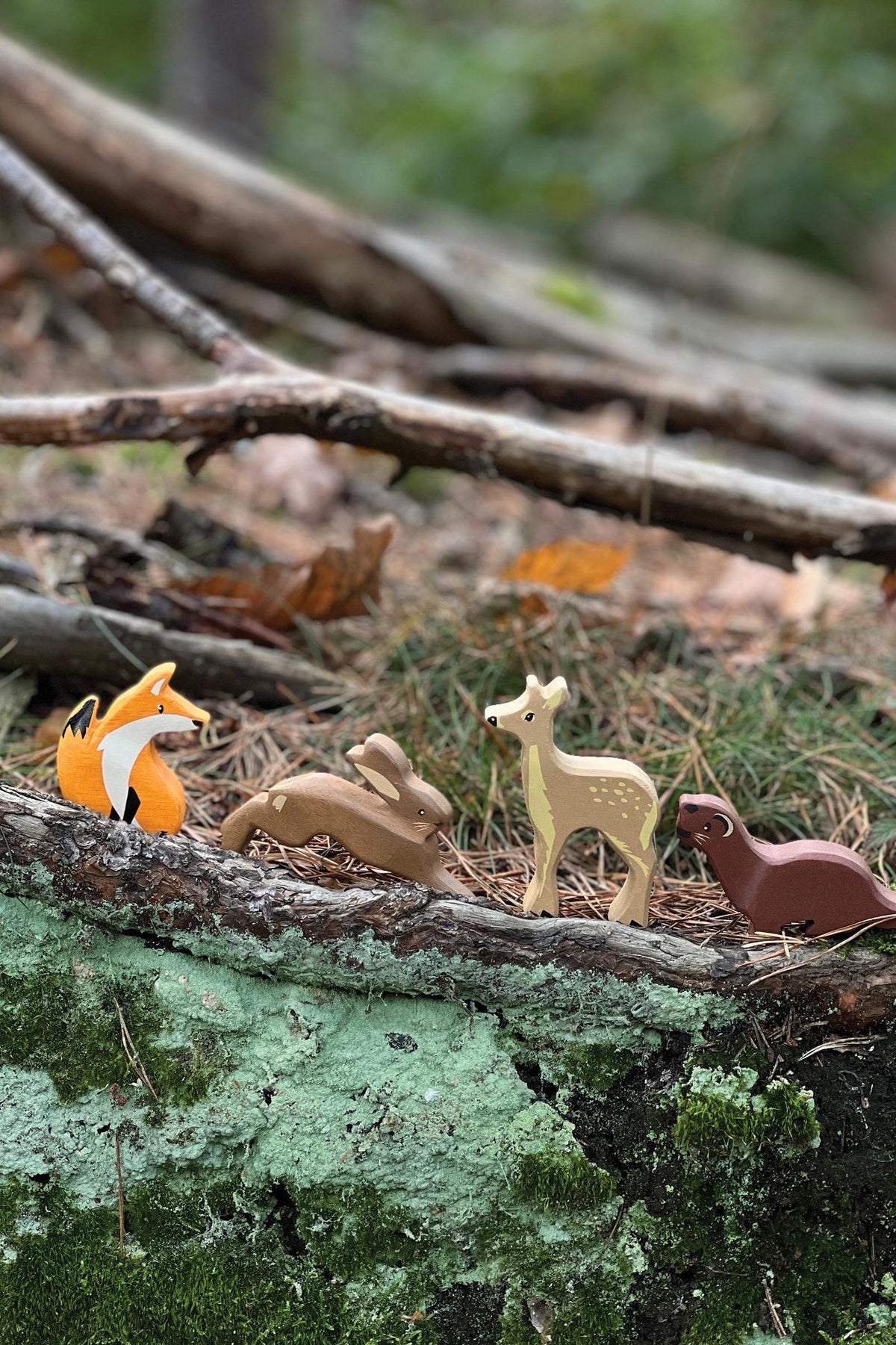 A collection of wooden woodland animal toys, including a fox, rabbit, deer, and weasel, is displayed on a natural forest floor among twigs and leaves, highlighting their eco-friendly design.