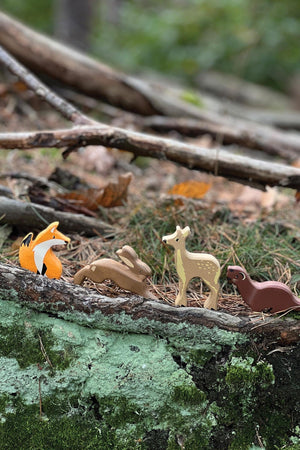 A collection of wooden woodland animal toys, including a fox, rabbit, deer, and weasel, is displayed on a natural forest floor among twigs and leaves, highlighting their eco-friendly design.