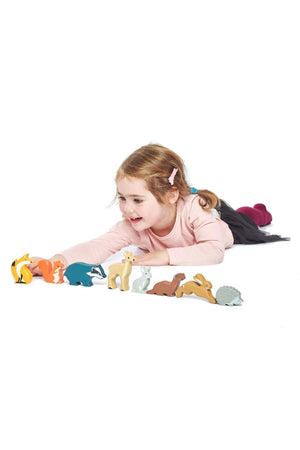 A young girl in a pink sweater lays on the floor, playing with colorful wooden toy animals from the Tender Leaf Woodland Animals collection, including a fox, deer, and hedgehog.